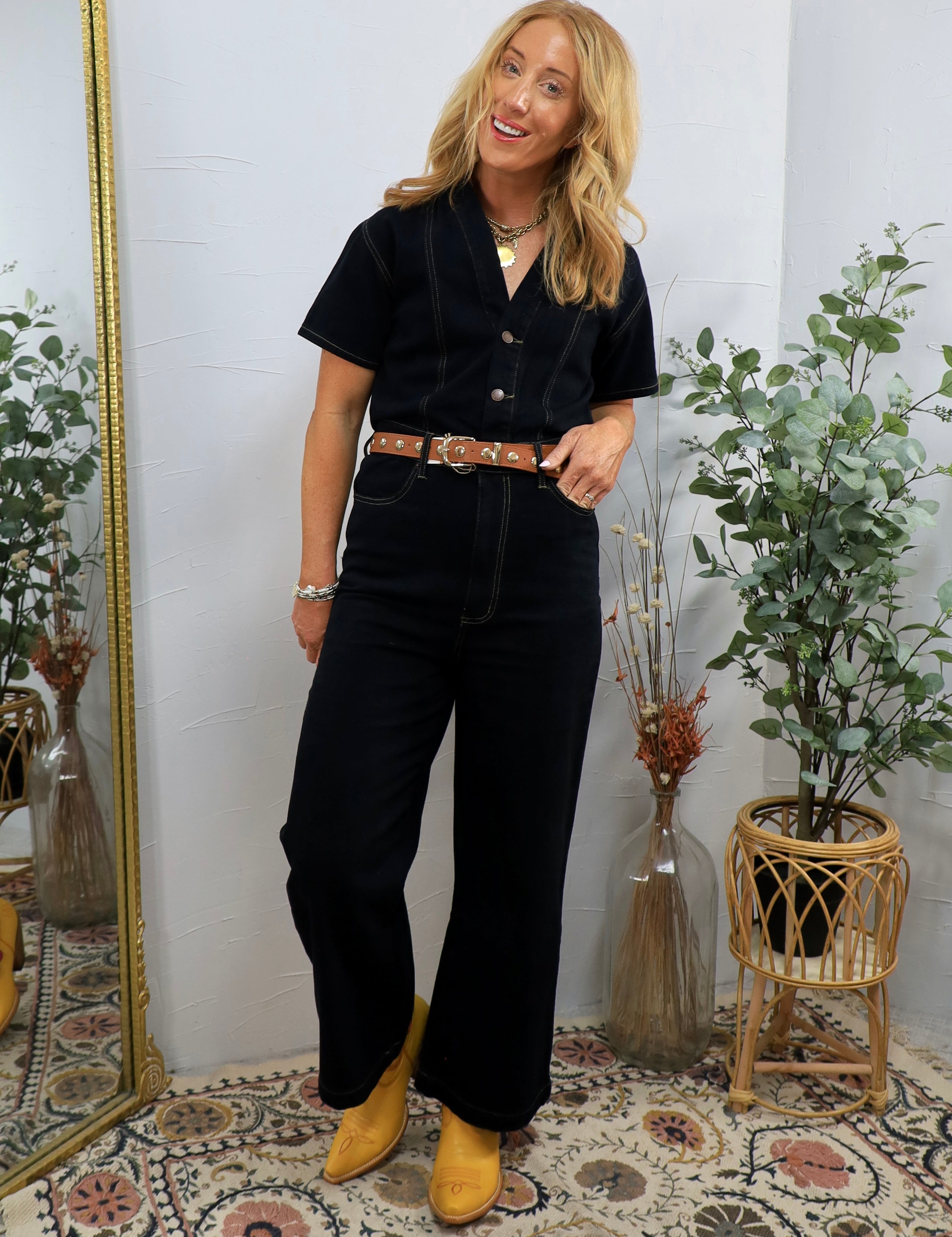 Woman wearing black short-sleeve denim coveralls with brown leather belt and yellow pointed-toe shoes, styled with layered ne