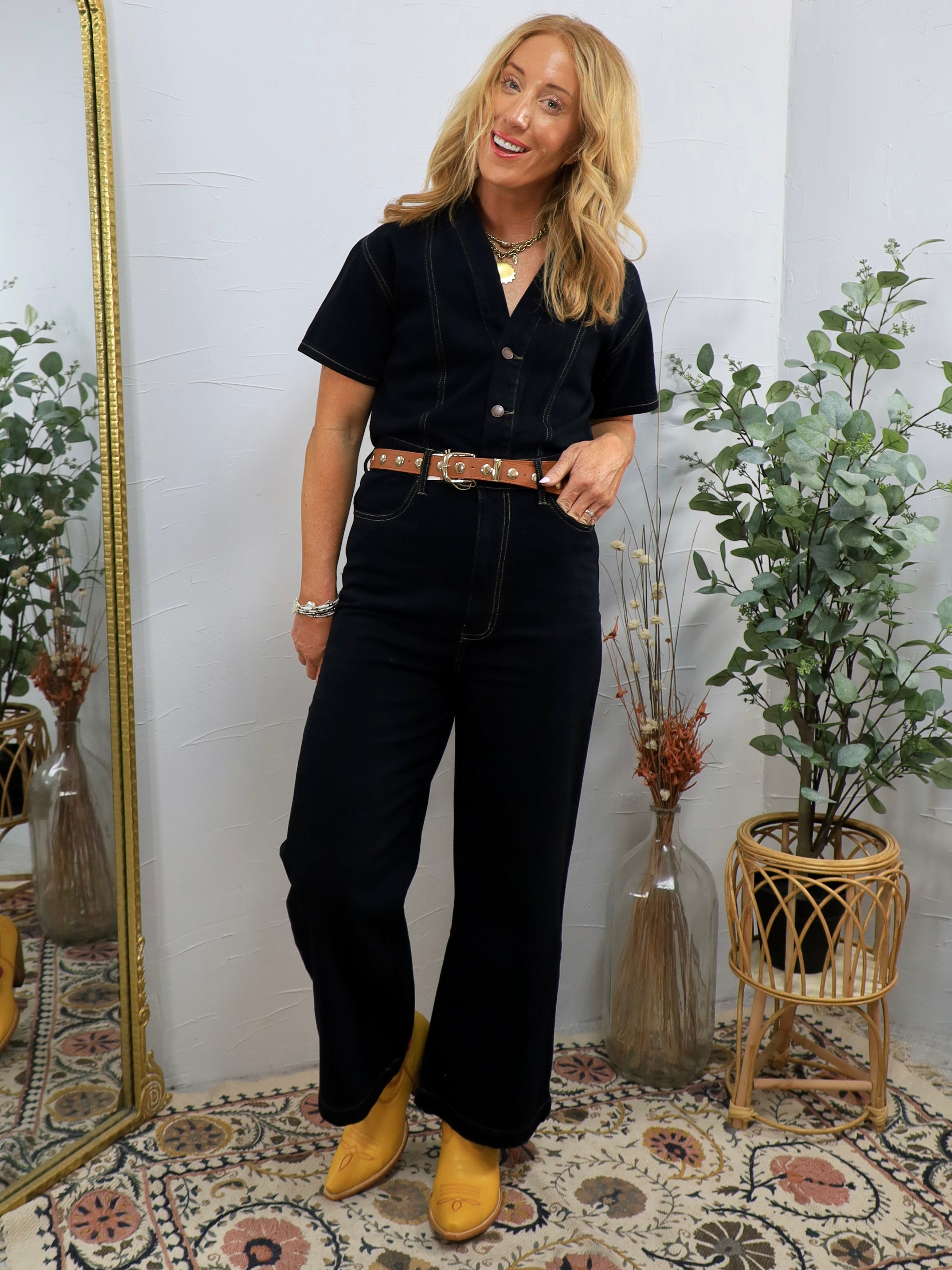 Woman wearing black short-sleeve denim coveralls with brown leather belt and yellow pointed-toe shoes, styled with layered ne