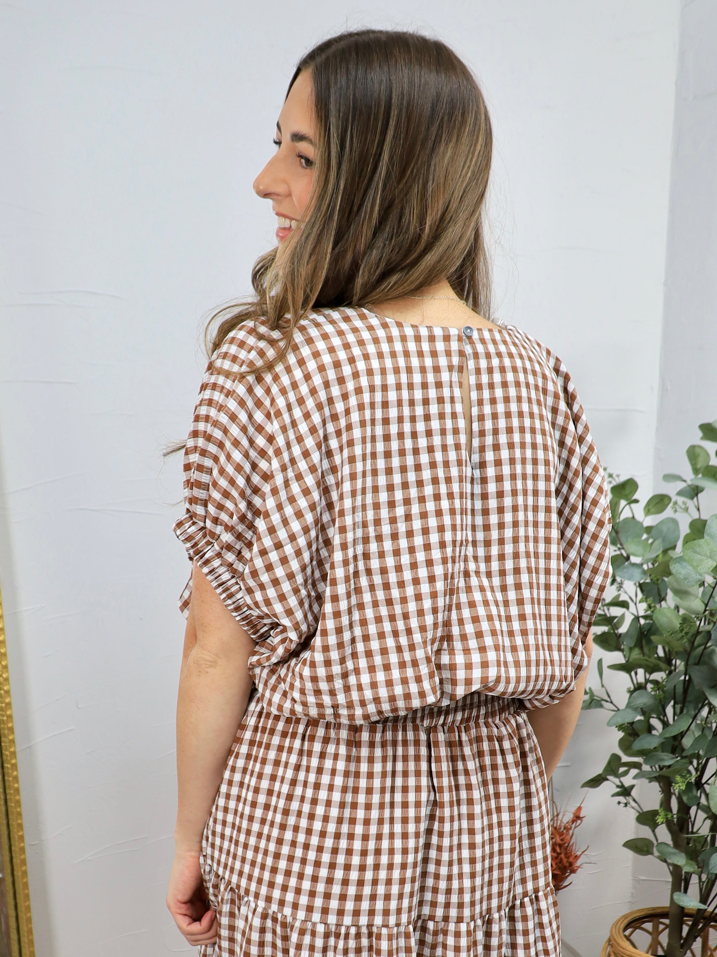 Woman wearing brown and white gingham checkered top with ruched short sleeves and elastic waistband, shown from behind.