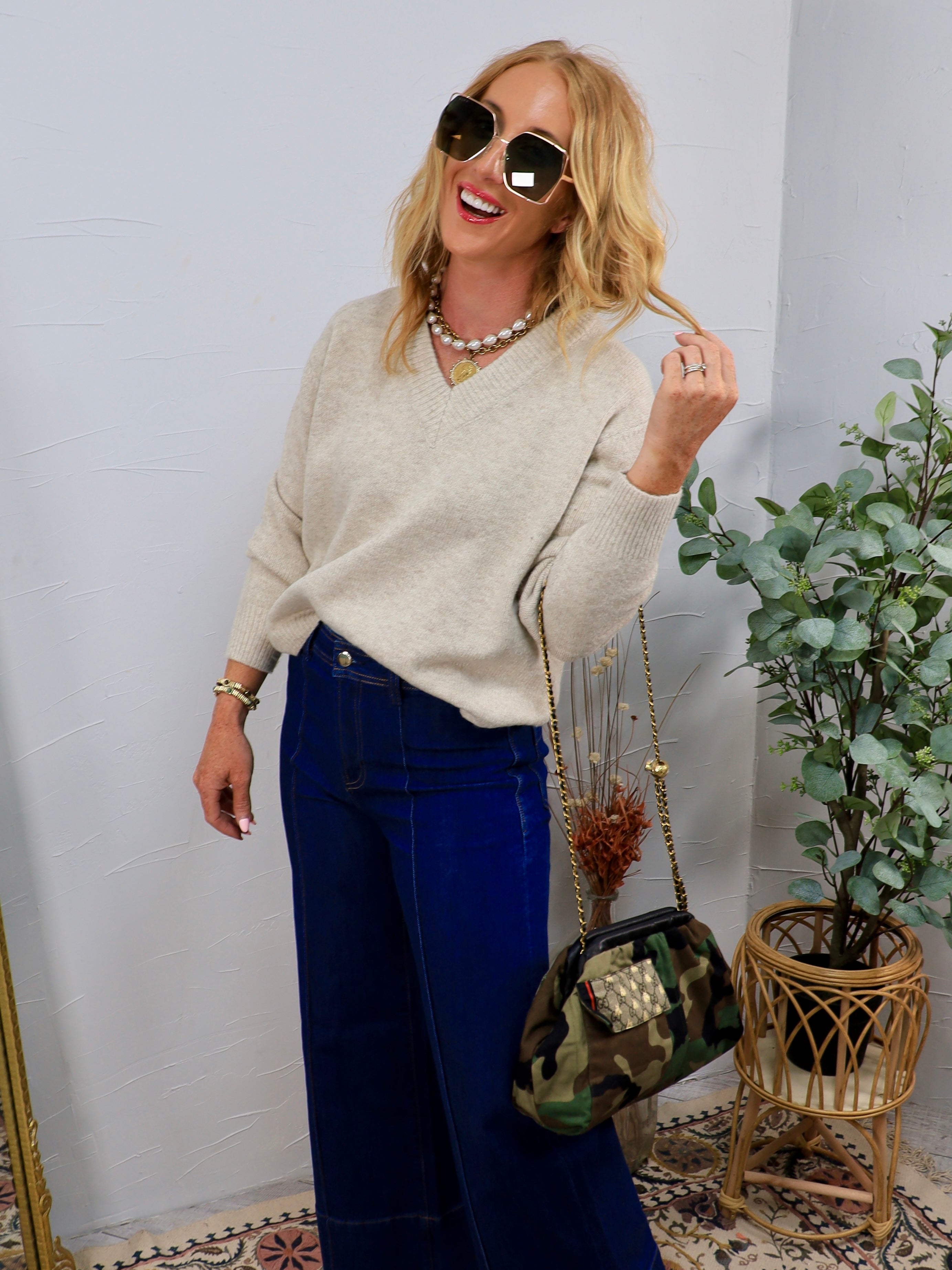 Woman wearing oatmeal V-neck oversized knit sweater with wide-leg dark denim jeans and layered gold jewelry