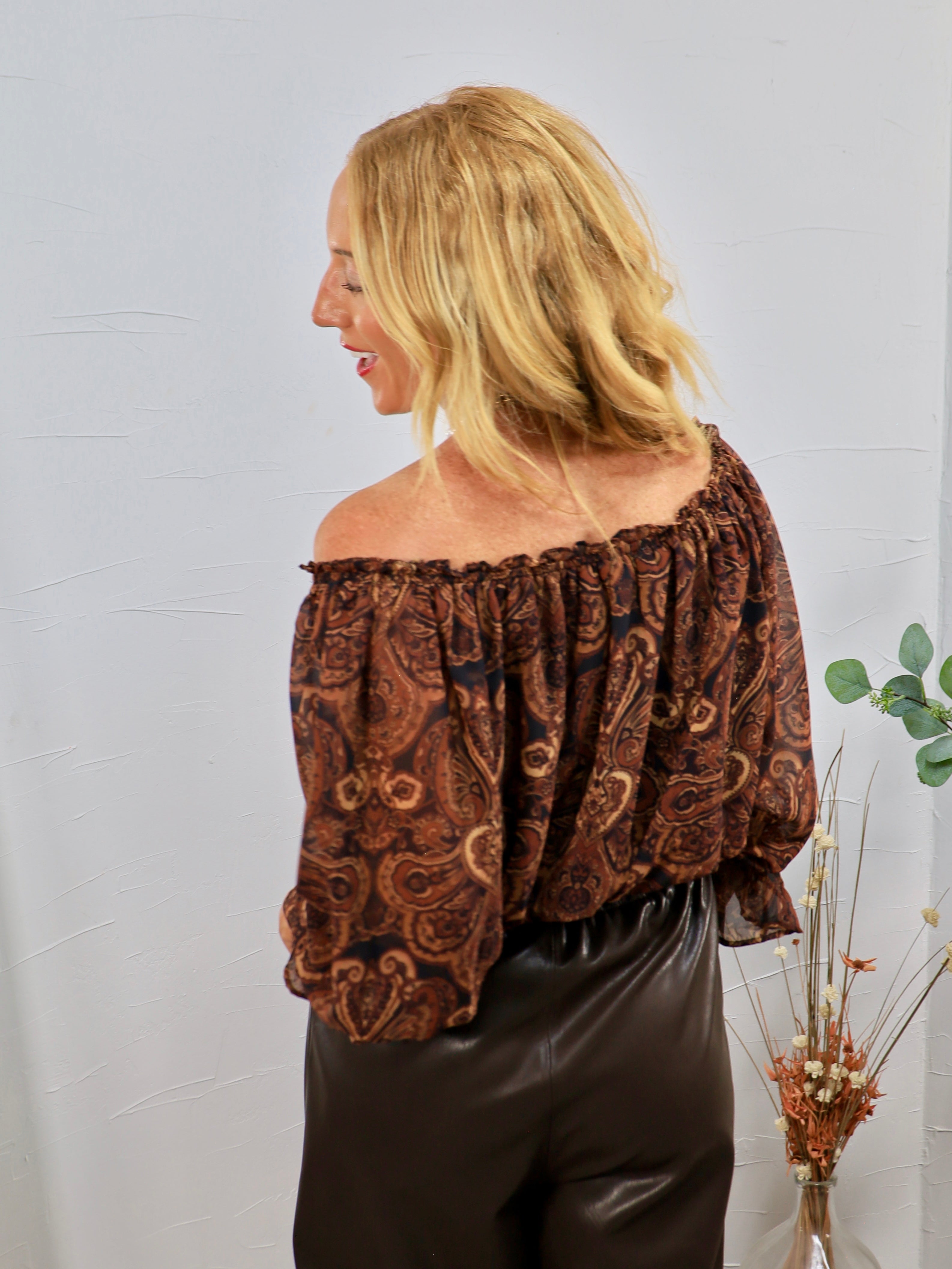 Brown and black paisley off-shoulder blouse with voluminous sleeves, styled with dark faux leather pants.