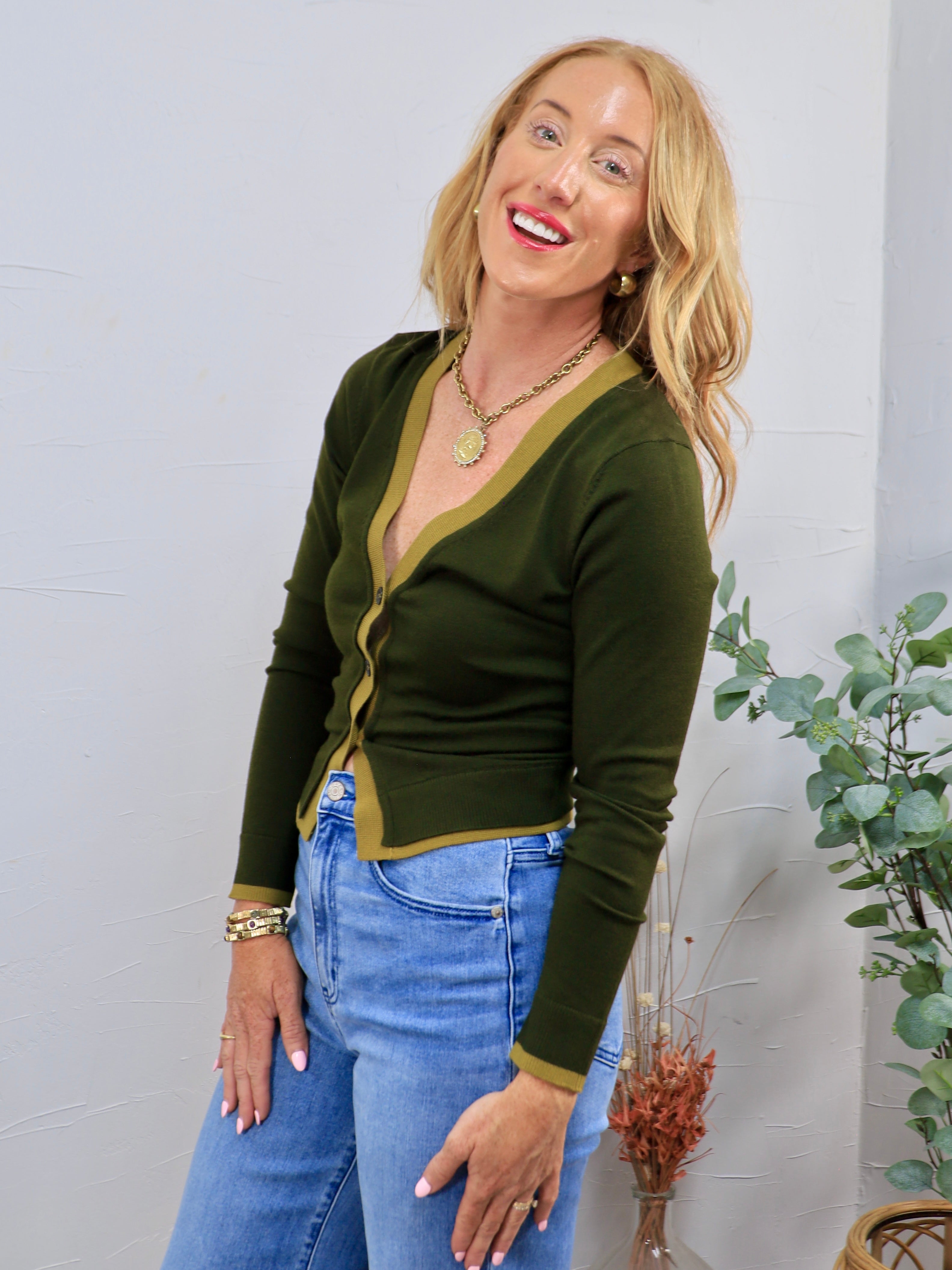 Woman wearing a cropped olive green button-front cardigan with contrast trim and blue jeans
