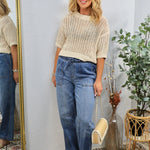 Woman wearing tan open-knit sweater top with blue wide-leg jeans and woven bag