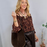 Woman wearing a dark brown paisley off-the-shoulder long sleeve blouse with a large brown studded tote bag