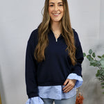 Woman wearing navy quarter-zip pullover with light blue layered shirt hem and ruffled cuffs with jeans