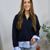 Woman wearing navy quarter-zip pullover with light blue layered shirt hem and ruffled cuffs with jeans