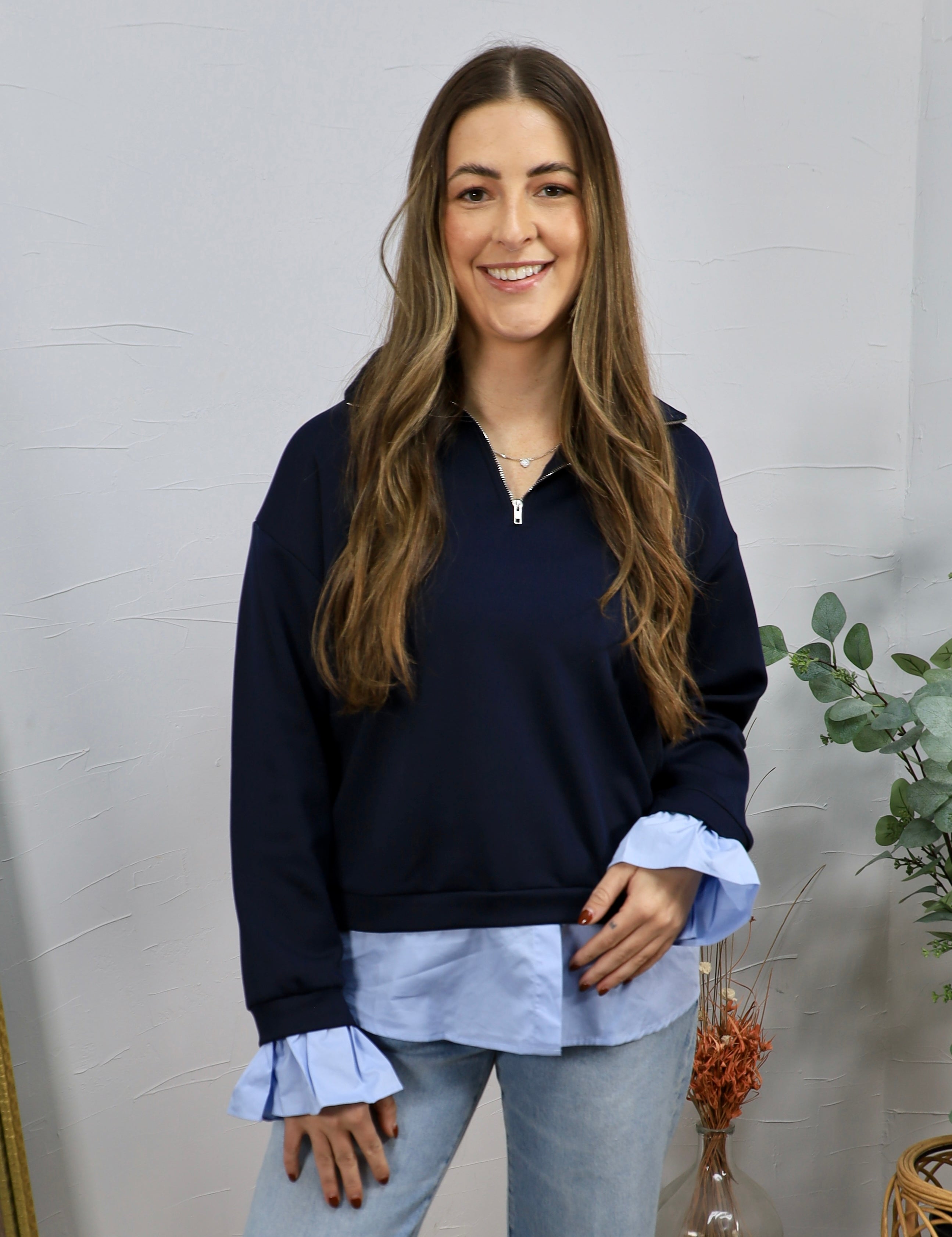 Woman wearing navy quarter-zip pullover with light blue layered shirt hem and ruffled cuffs with jeans