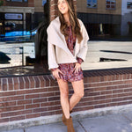 Woman wearing a cropped cream shawl-collar jacket over a floral mini dress with tan ankle booties outdoors