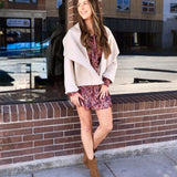 Woman wearing a cropped cream shawl-collar jacket over a floral mini dress with tan ankle booties outdoors