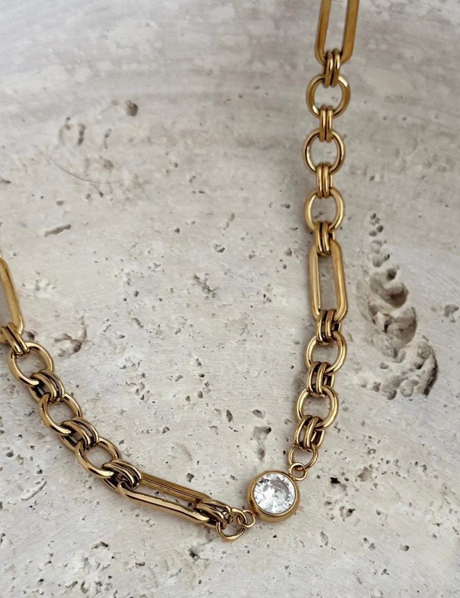 Gold mixed-link chain necklace with a bezel-set clear crystal charm on stone surface