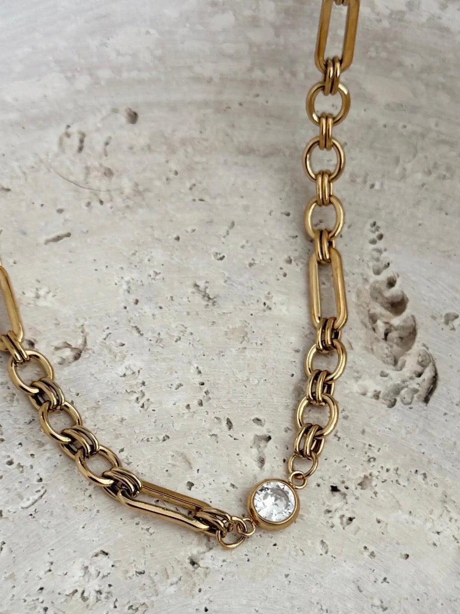 Gold mixed-link chain necklace with a bezel-set clear crystal charm on stone surface