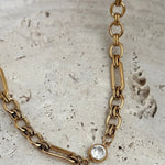 Gold mixed-link chain necklace with a bezel-set clear crystal charm on stone surface