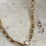 Gold mixed-link chain necklace with a bezel-set clear crystal charm on stone surface