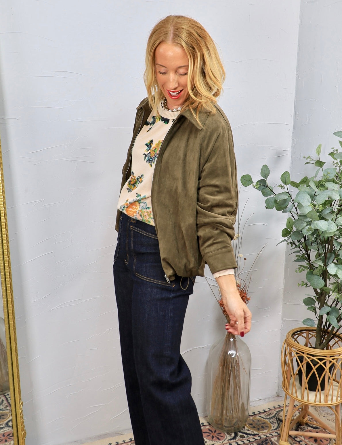 Woman wearing olive green ruched suede bomber jacket over floral sweater with dark wide-leg jeans
