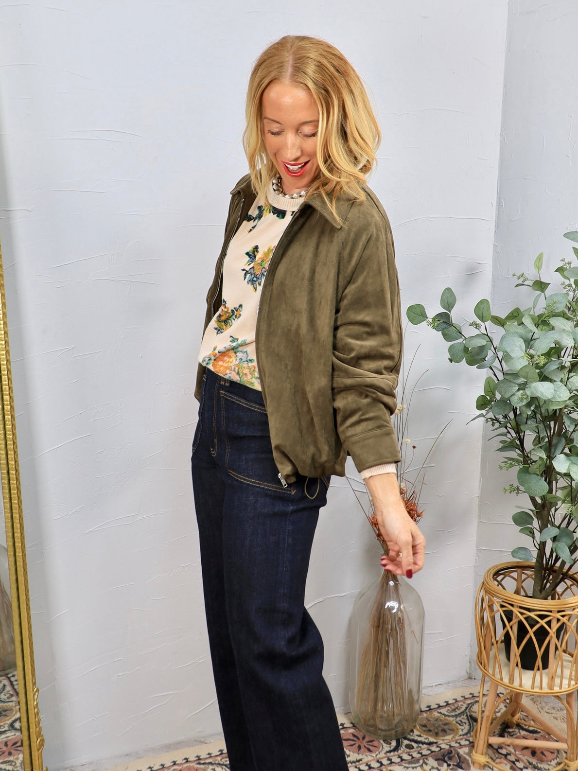 Woman wearing olive green ruched suede bomber jacket over floral sweater with dark wide-leg jeans