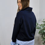 Navy blue pullover sweatshirt layered over light blue collared shirt, styled with light wash jeans, back view.