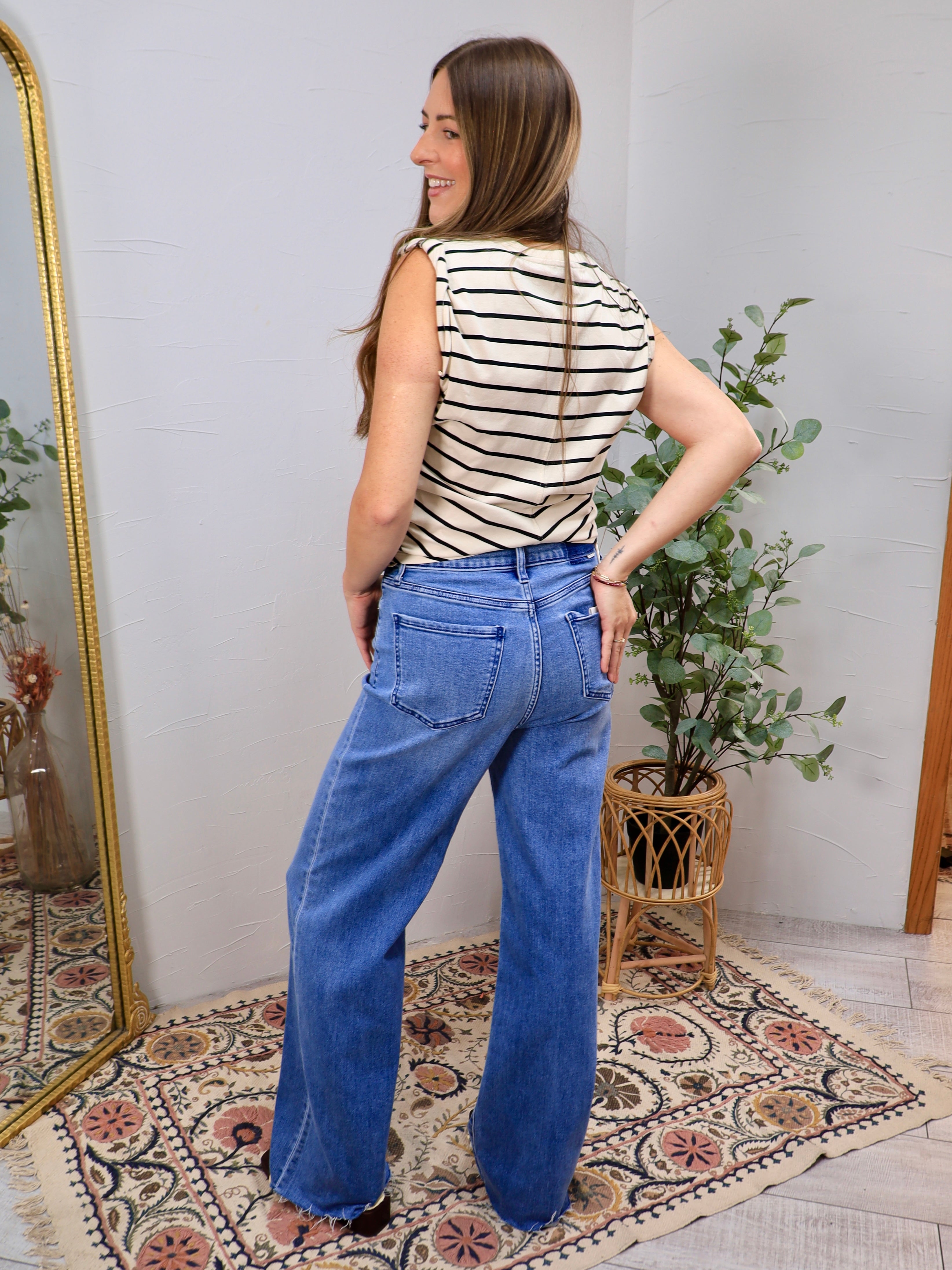 Wide-leg medium wash blue denim jeans with raw hem, styled with striped sleeveless top, back view.