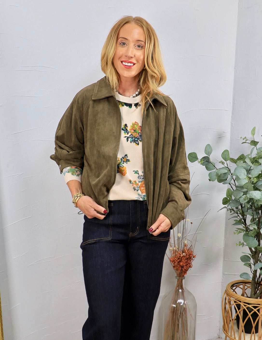 Olive green suede zip-up bomber jacket with ruched sleeves, worn open over a floral sweater with dark jeans.