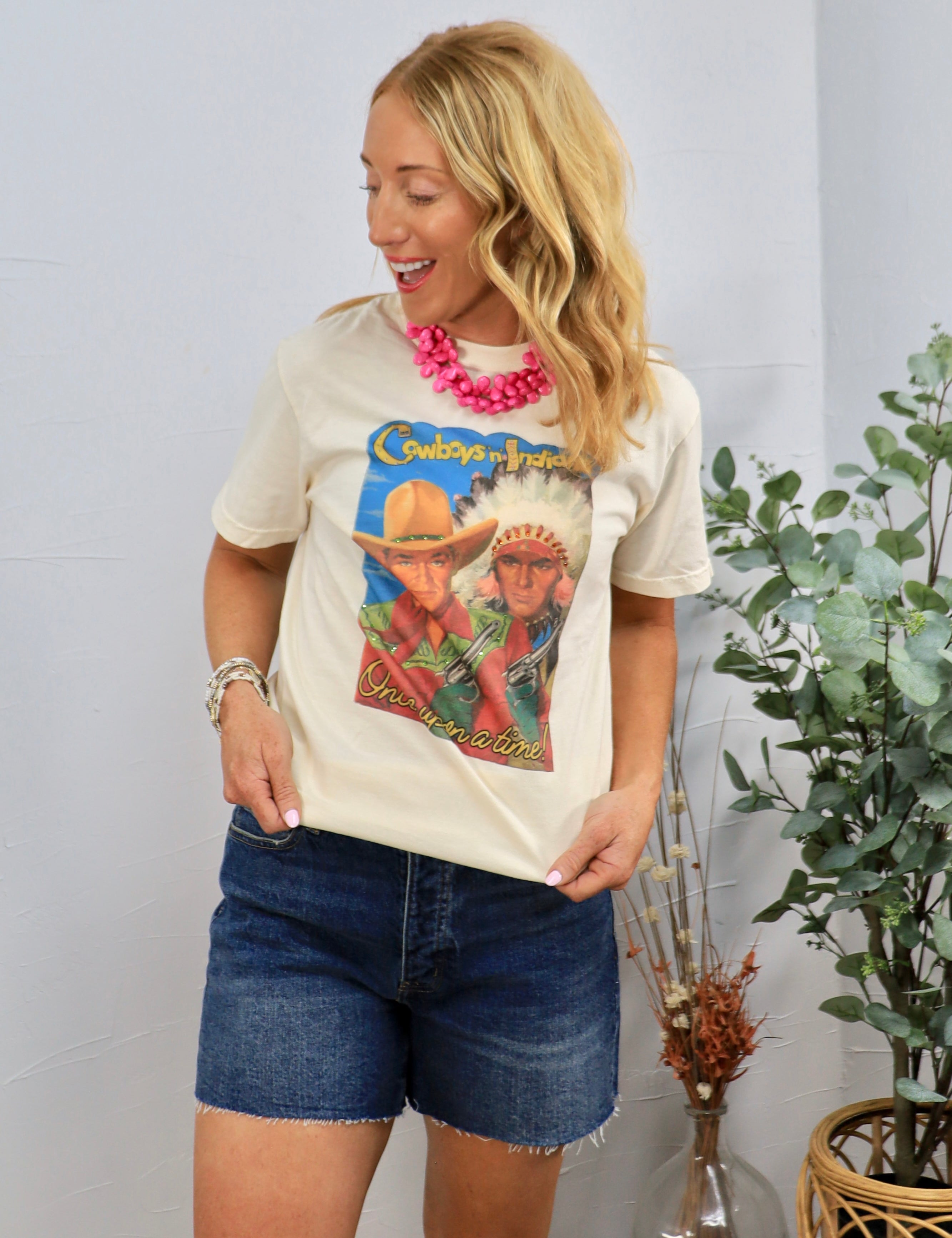 Woman wearing cream Cowboys and Indians graphic tee with pink statement necklace and denim shorts in casual styling.