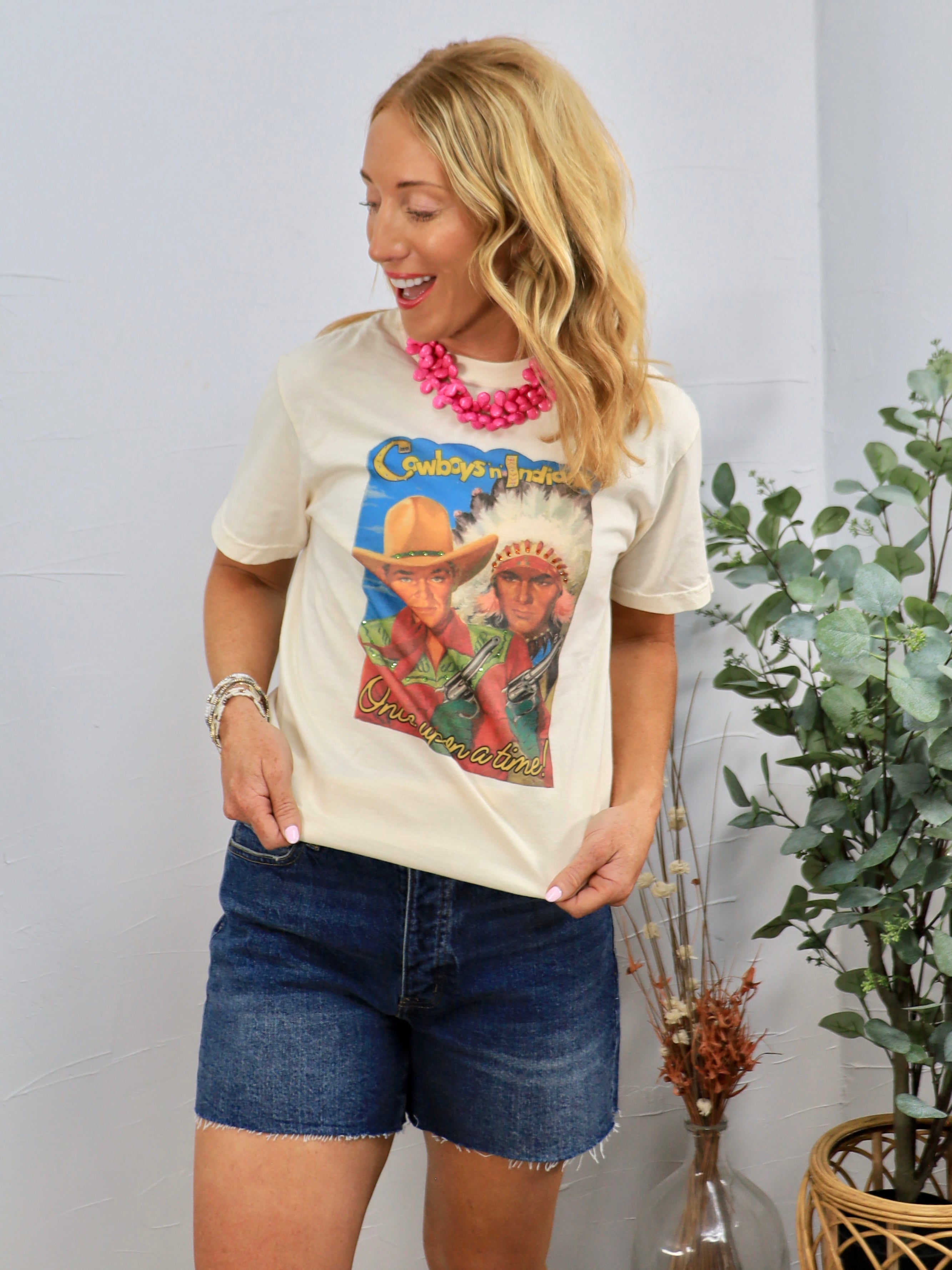 Woman wearing cream Cowboys and Indians graphic tee with pink statement necklace and denim shorts in casual styling.