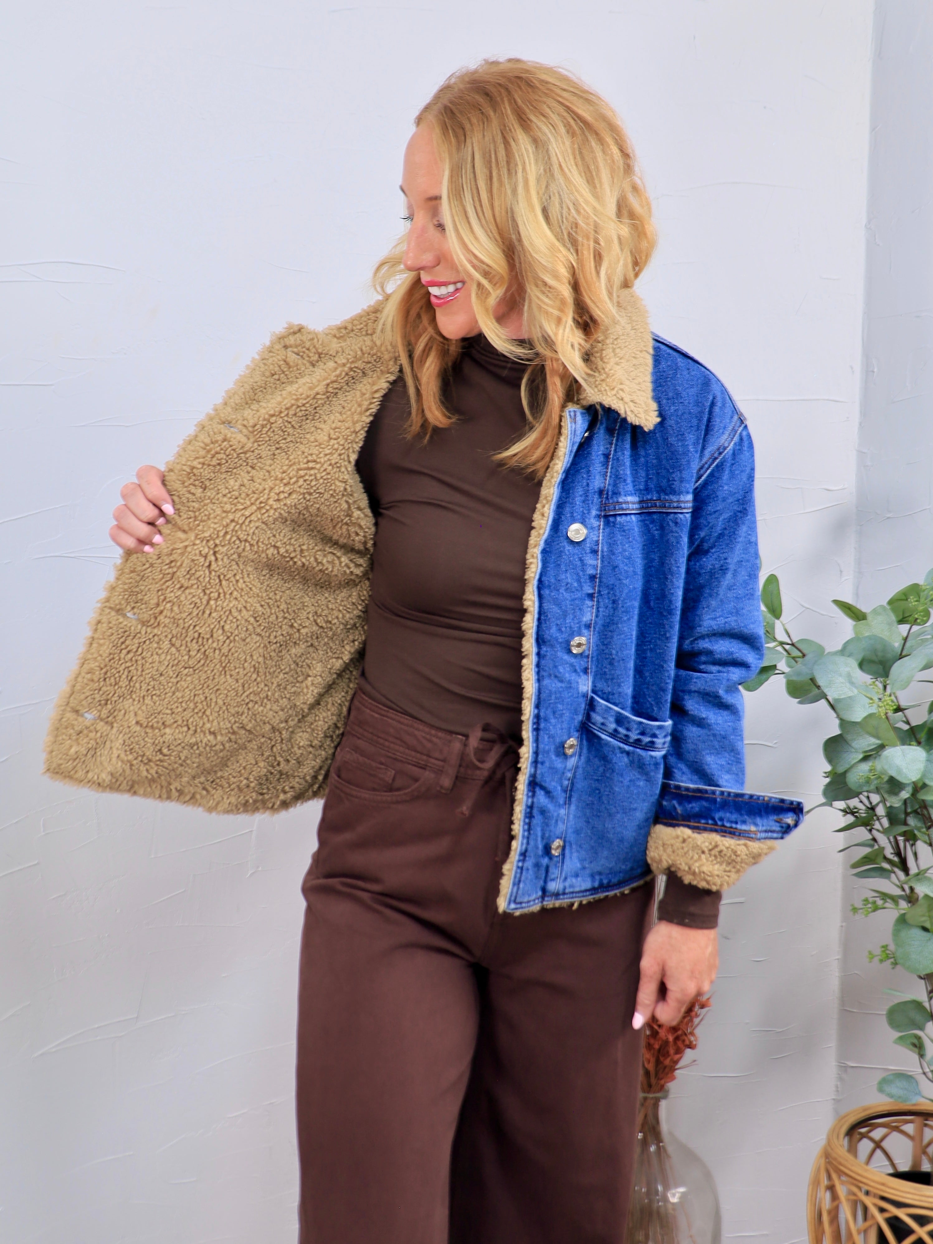 Blue denim jacket with tan sherpa teddy lining and collar, shown open, styled over brown turtleneck and pants.