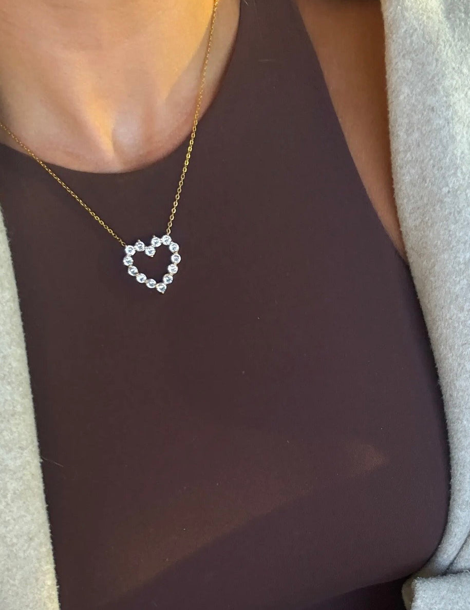 Delicate gold chain necklace with open heart pendant made of sparkling round crystal stones worn over a brown top.