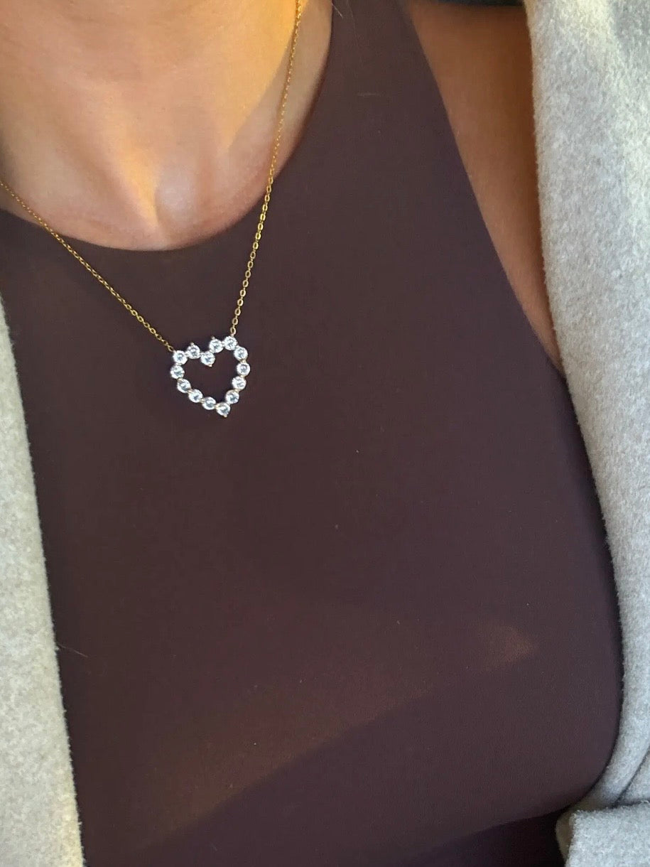 Delicate gold chain necklace with open heart pendant made of sparkling round crystal stones worn over a brown top.