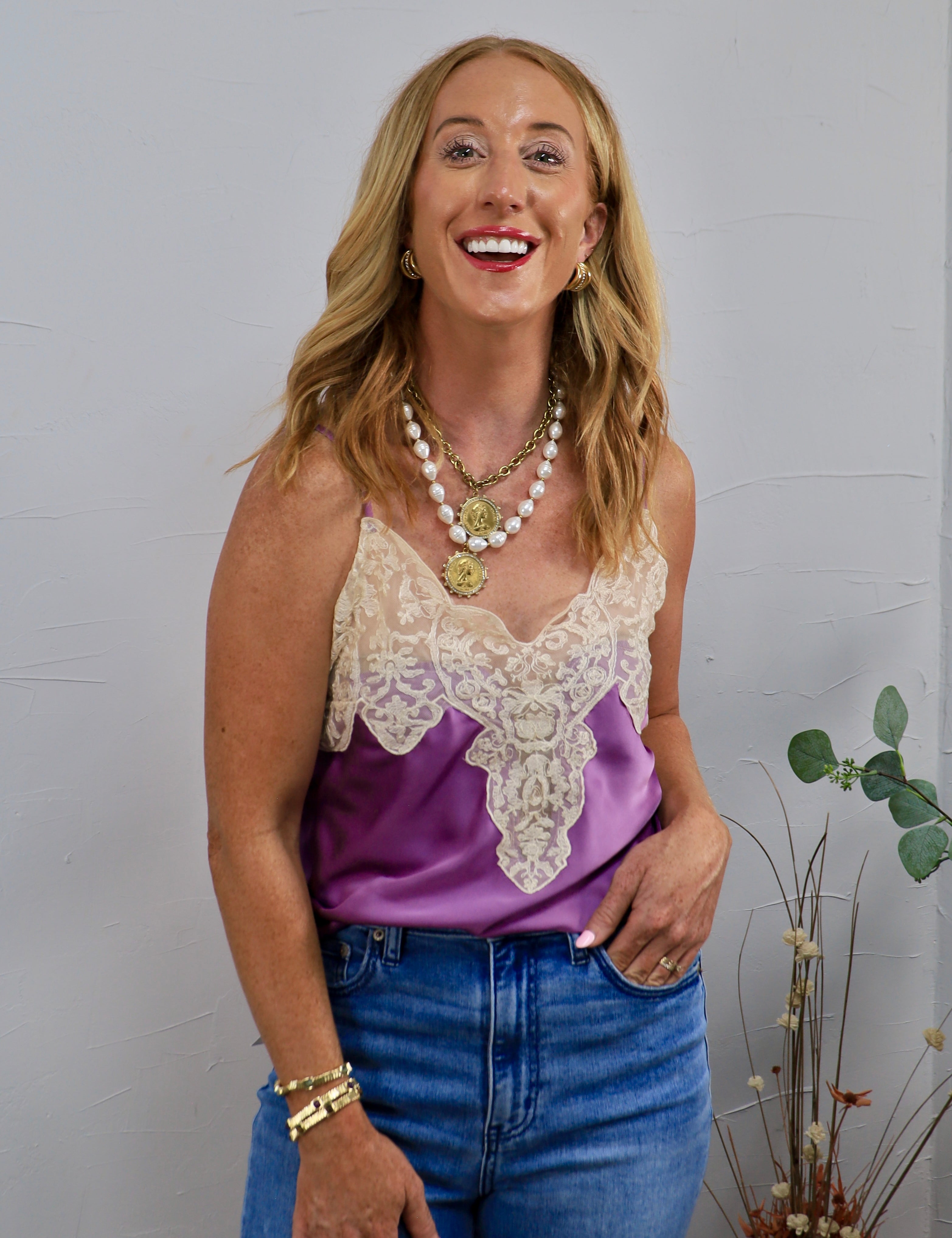 Woman wearing a purple satin spaghetti strap cami with cream lace neckline detailing and blue jeans