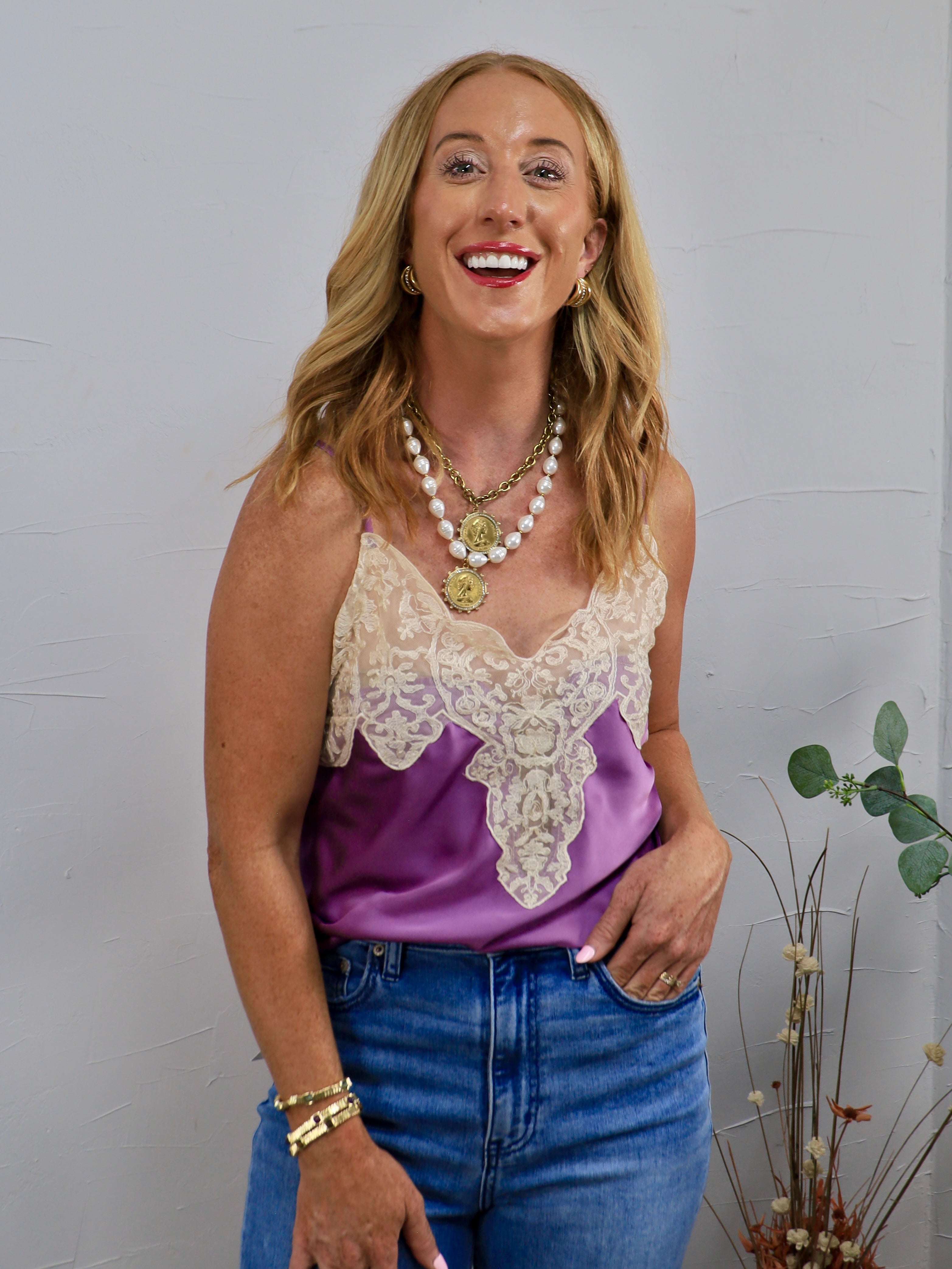 Woman wearing a purple satin spaghetti strap cami with cream lace neckline detailing and blue jeans