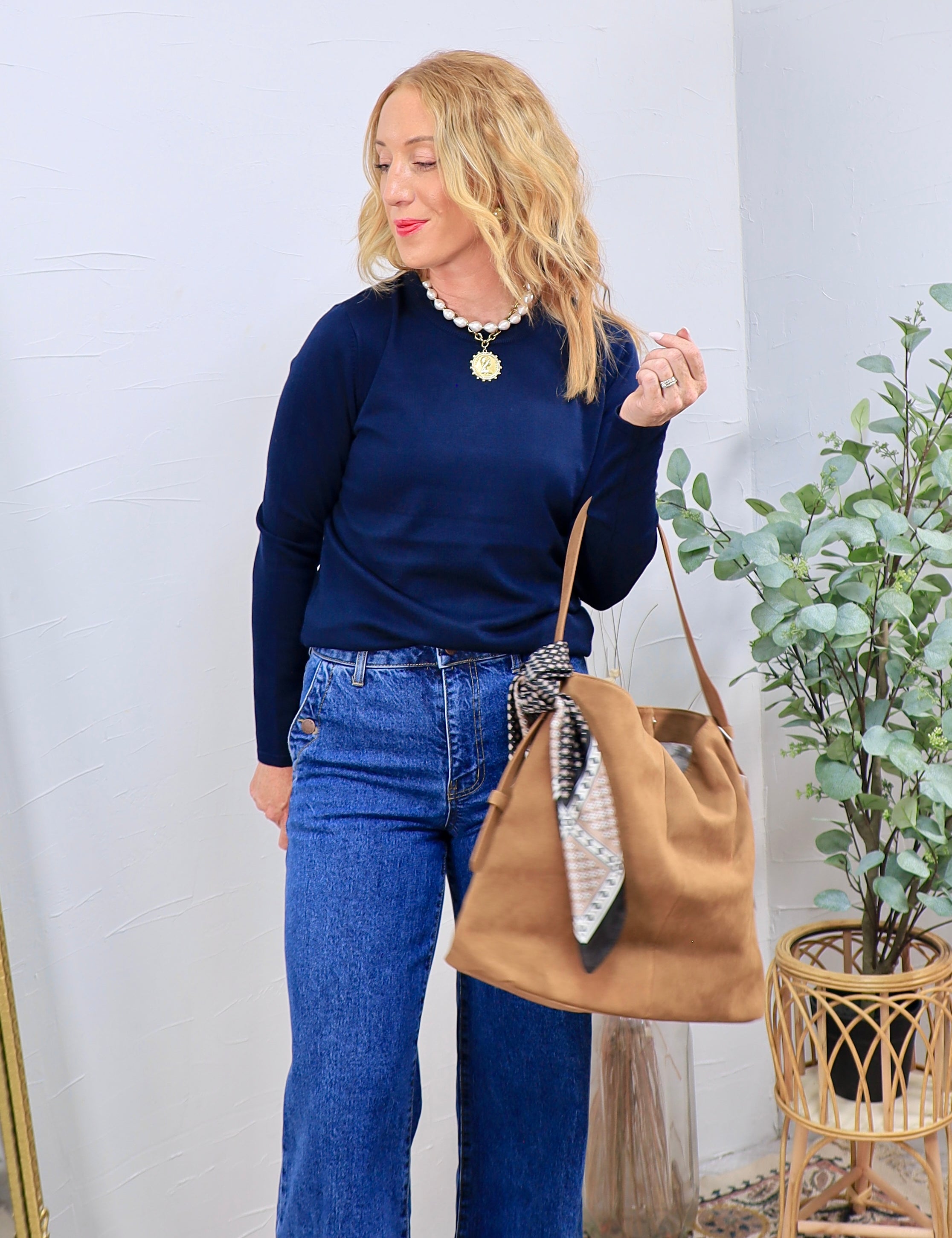 Woman in navy long sleeve sweater, wide-leg jeans, pearl necklace, and camel tote bag