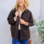 Dark brown oversized utility jacket with snap buttons, worn open over cream blouse and blue jeans.
