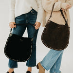 Two women holding large black and brown suede studded hobo tote bags with leather straps