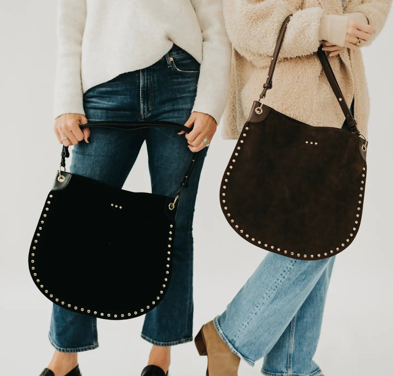 Two women holding large black and brown suede studded hobo tote bags with leather straps