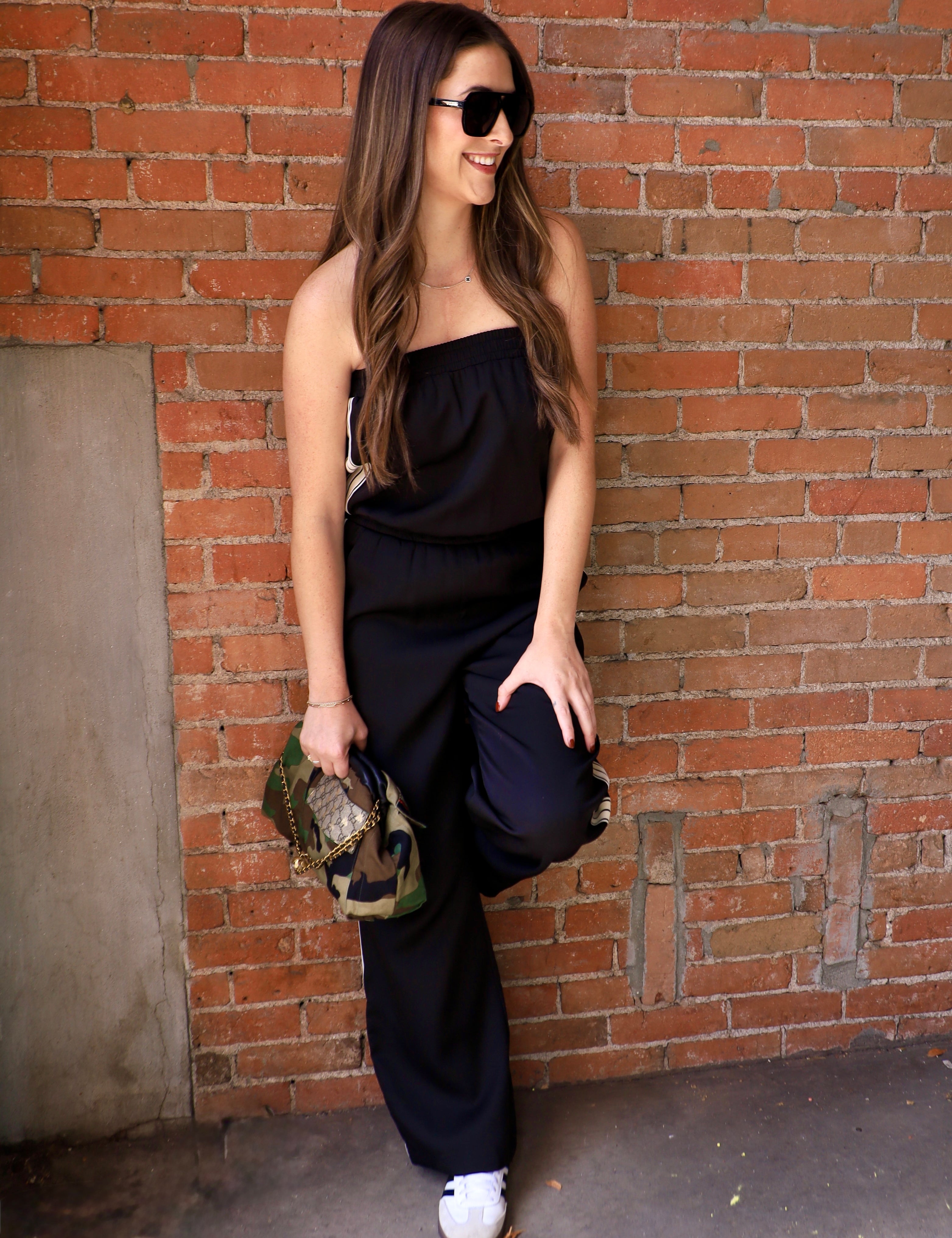 Woman wearing a black strapless wide-leg jumpsuit with white sneakers and camo bag against brick wall