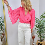 Hot pink wrap blouse with dramatic pleated bell sleeves, styled with white wide-leg jeans.