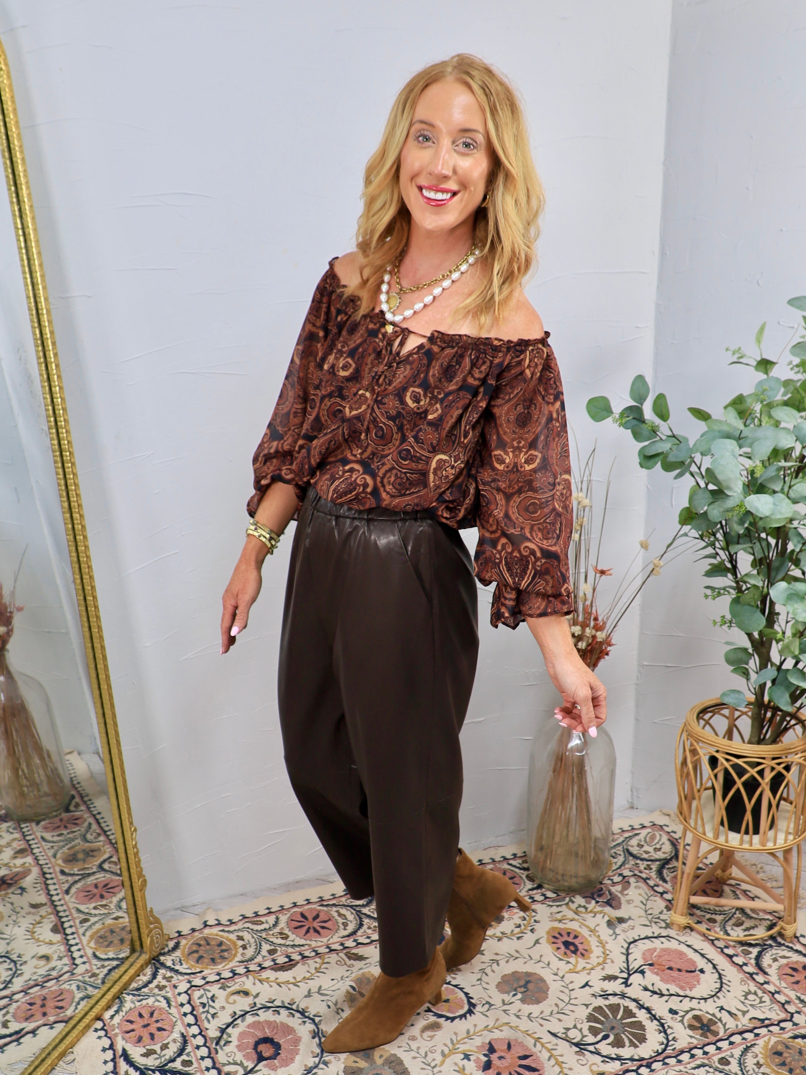 Dark brown faux leather tapered pants styled with paisley off-shoulder top and tan ankle boots.