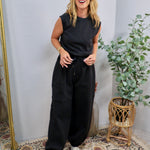 Black sleeveless jumpsuit with drawstring waist, padded shoulders, and jogger-style tapered legs, styled with silver sneakers