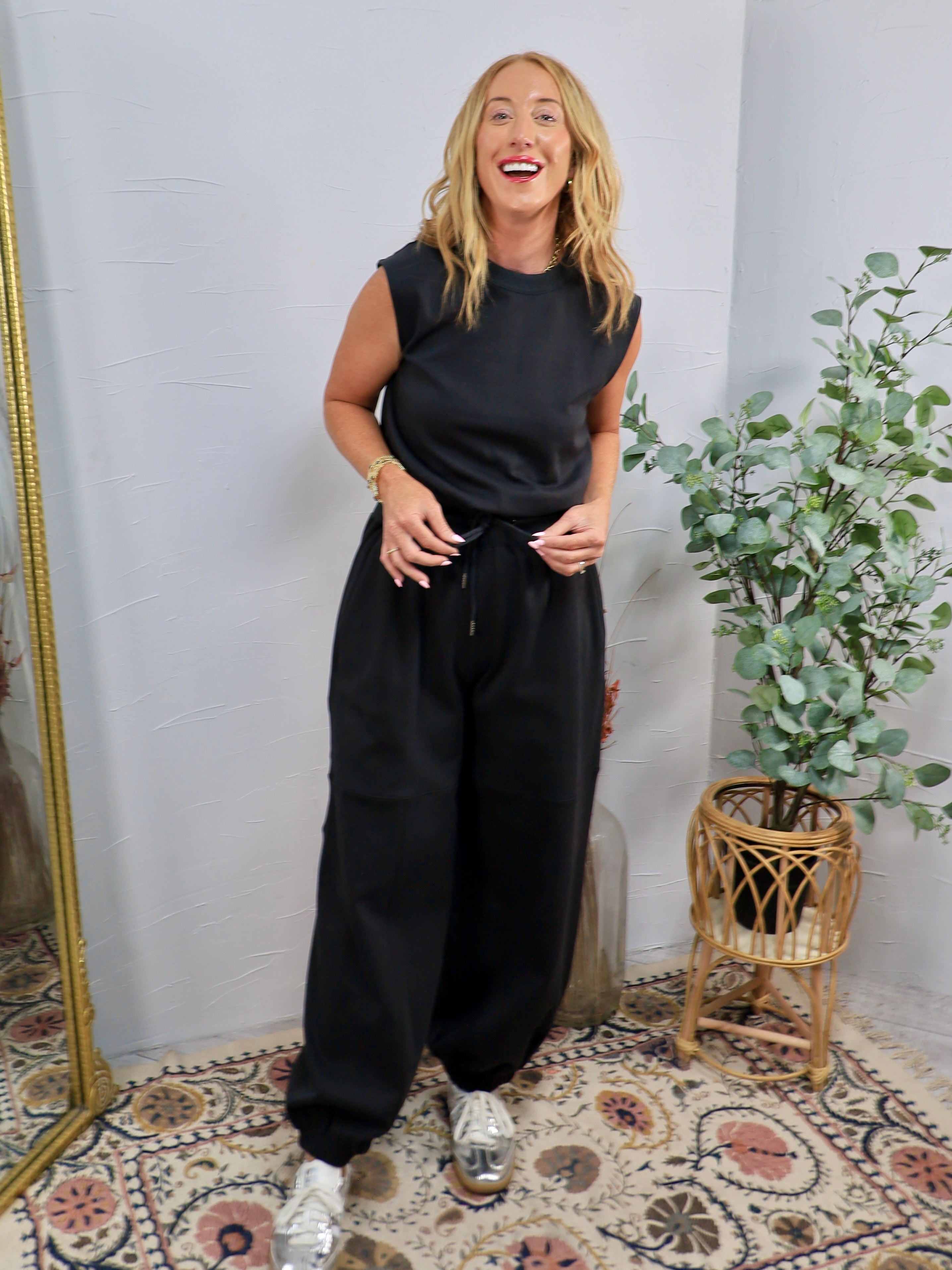 Black sleeveless jumpsuit with drawstring waist, padded shoulders, and jogger-style tapered legs, styled with silver sneakers