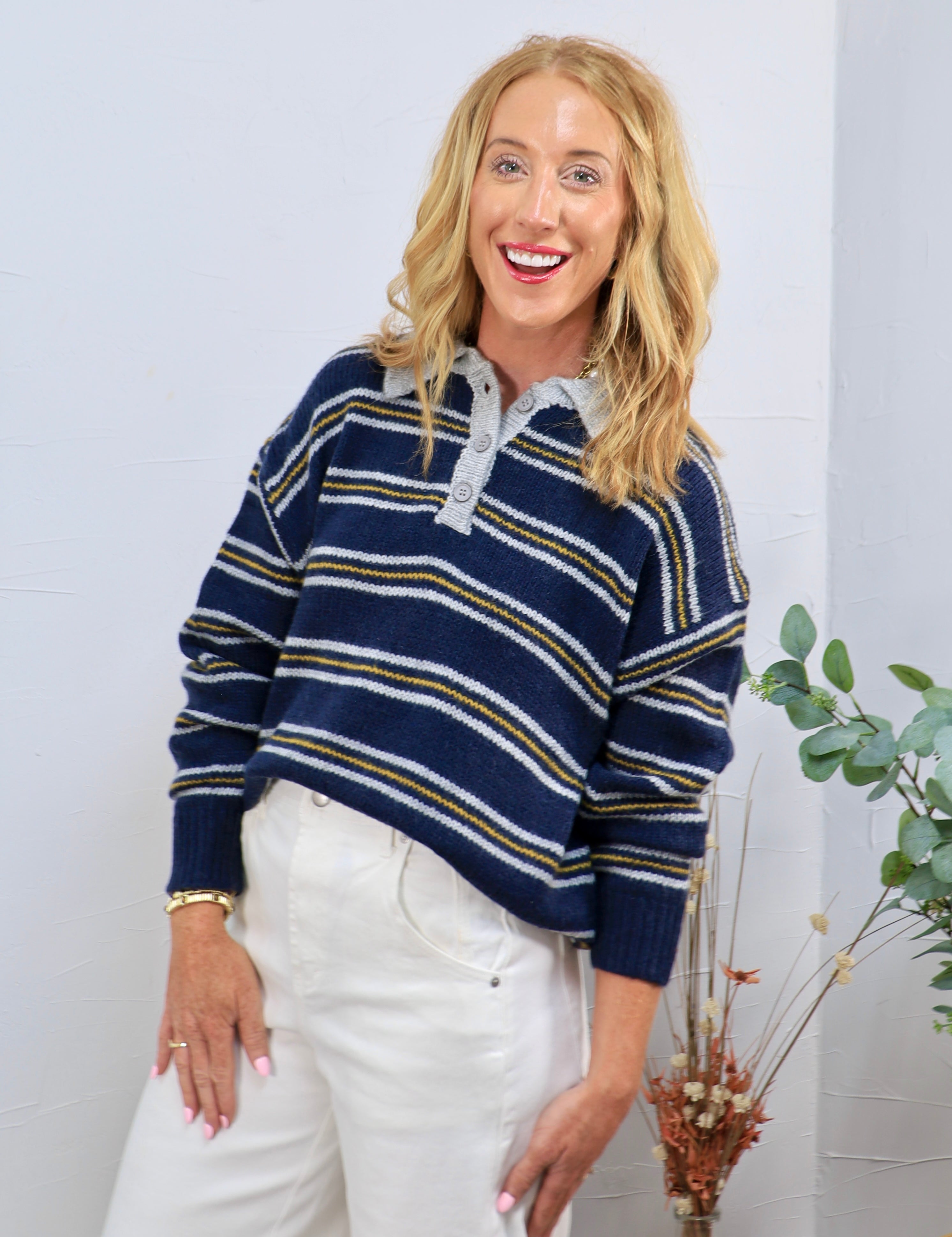 Navy blue striped knit polo sweater with gold and white stripes, gray collar, and button placket, styled with white trousers.