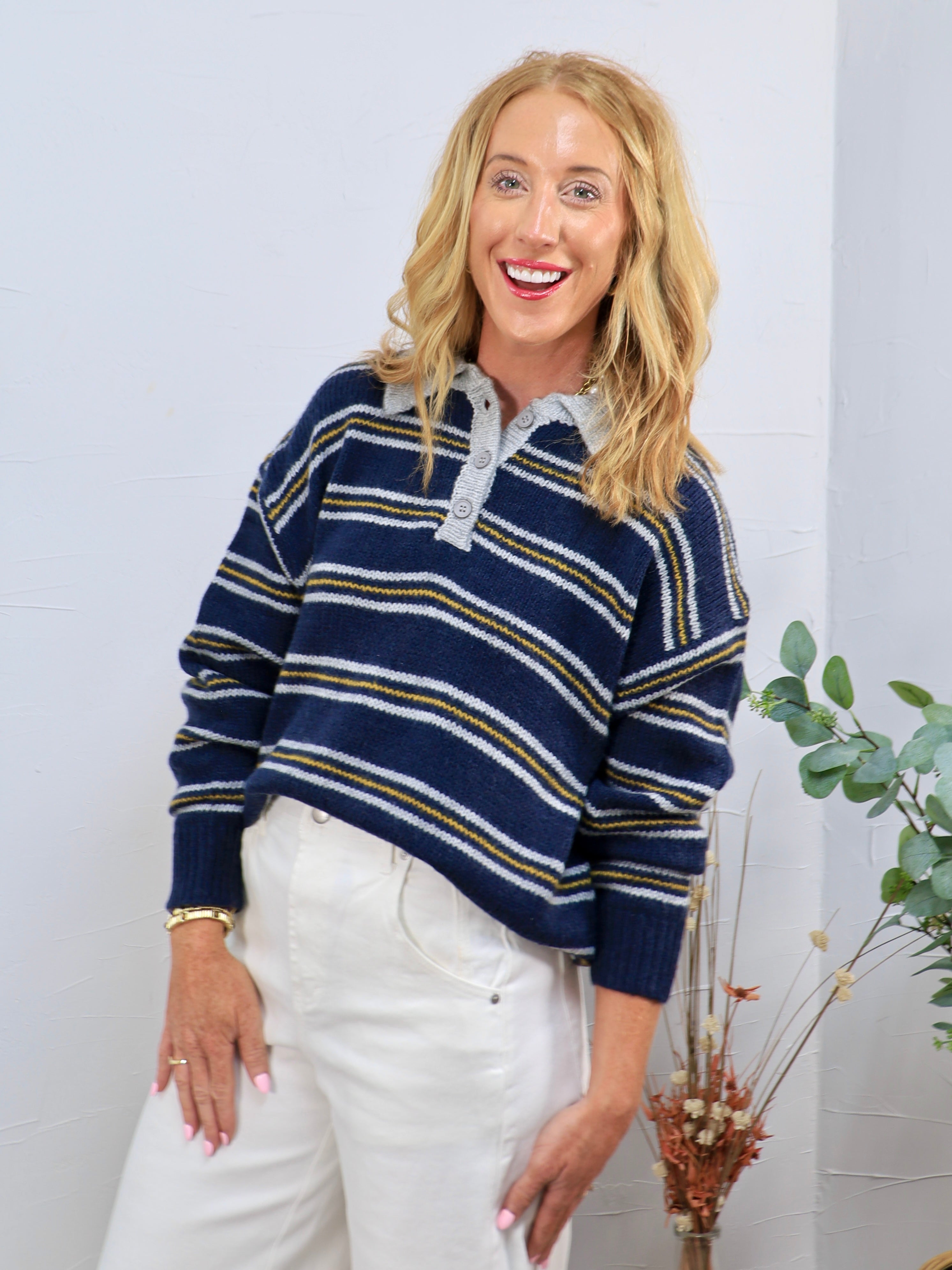 Navy blue striped knit polo sweater with gold and white stripes, gray collar, and button placket, styled with white trousers.