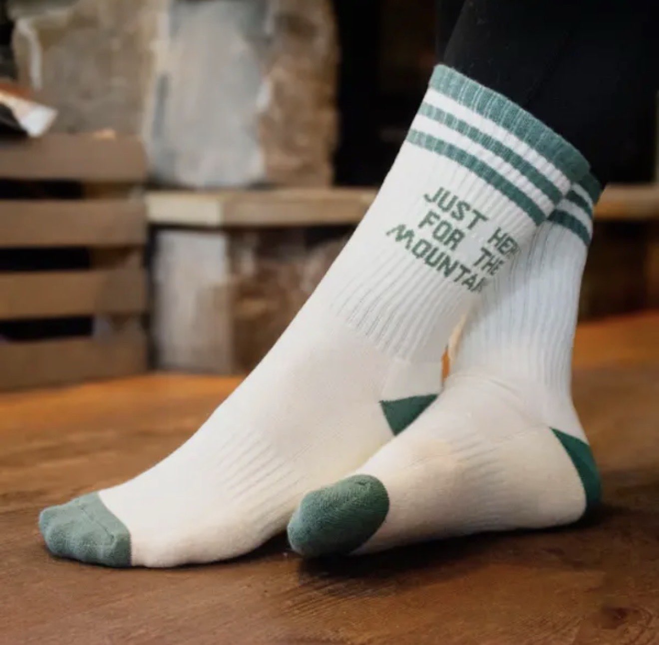 White crew socks with green accents and 'Just For The Mountains' text displayed on wooden surface