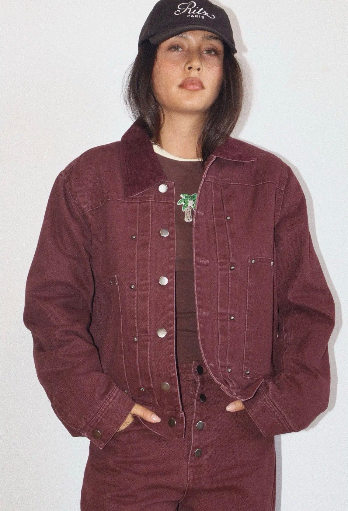 Oversized burgundy denim bomber jacket with silver snap buttons, corduroy collar, and palm tree brooch detail.